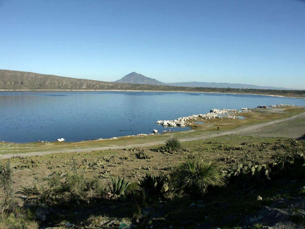 100227_Alchichica 012.jpg - Laguna Alchichica. Pověstmi opředená vulkanická laguna (jezero uvnitř starého vulkanického kráteru, ale tady říkají všichni laguna) se slanou vodou v těsné blízkosti stejnojmenné vesnice. Vesnicí prochází státní silnice spojující Perote a Acatzingo, takže spíše připomíná shluk dílen a garáží. Někteří lidé věří, že tato laguna je místo, kde občas přistávají vesmírné lodě mimozemšťanů. Prý viděli světla, která kroužila na nebi okolo středu nad lagunou, potom se shlukla v jeden bod a zmizela pod hladinou přímo v jejím středu. Údajně se také ještě nepodařilo změřit hloubku laguny. Nevím, co je na tom pravdy, z morfologického hlediska leží laguna ve vulkanickém kráteru s výrazně asymetrickým okrajem, který je na západní straně značně vyšší.